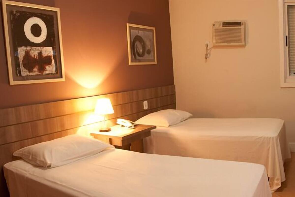 A hotel room with two beds, a bedside table with a lamp, a TV, and a window with blinds.