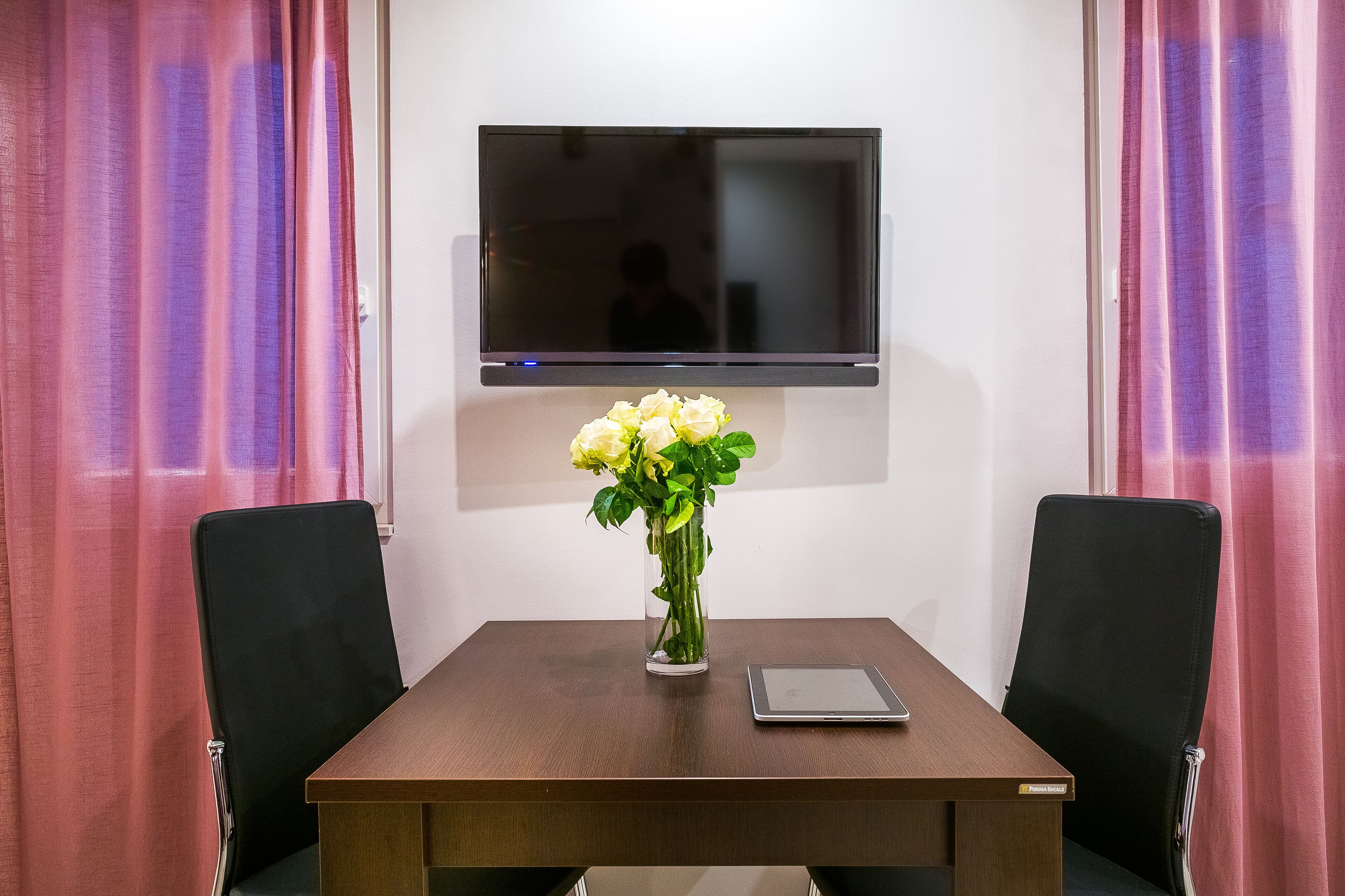 Una sala conferenze con tavolo in legno, due sedie nere, un vaso in vetro con fiori bianchi, un tablet e una televisione a schermo piatto montata a parete.