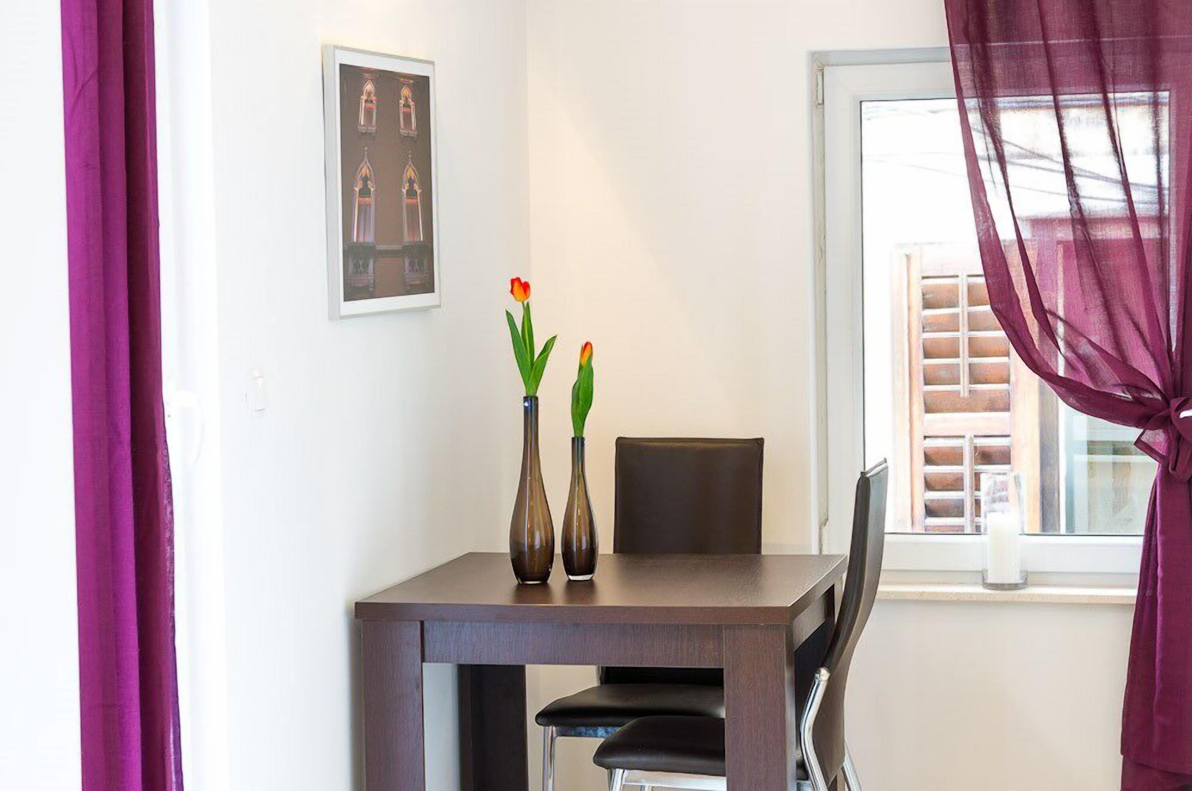 A dining area with a table, chairs, two vases with flowers, a framed picture on the wall, and a window with curtains.