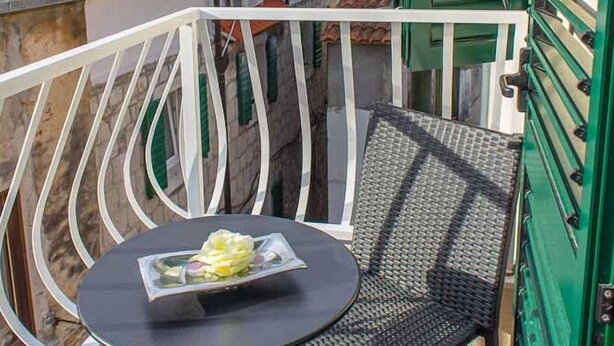 Un balcón con una mesa redonda y dos sillas, una verja verde, y vistas a edificios vecinos.