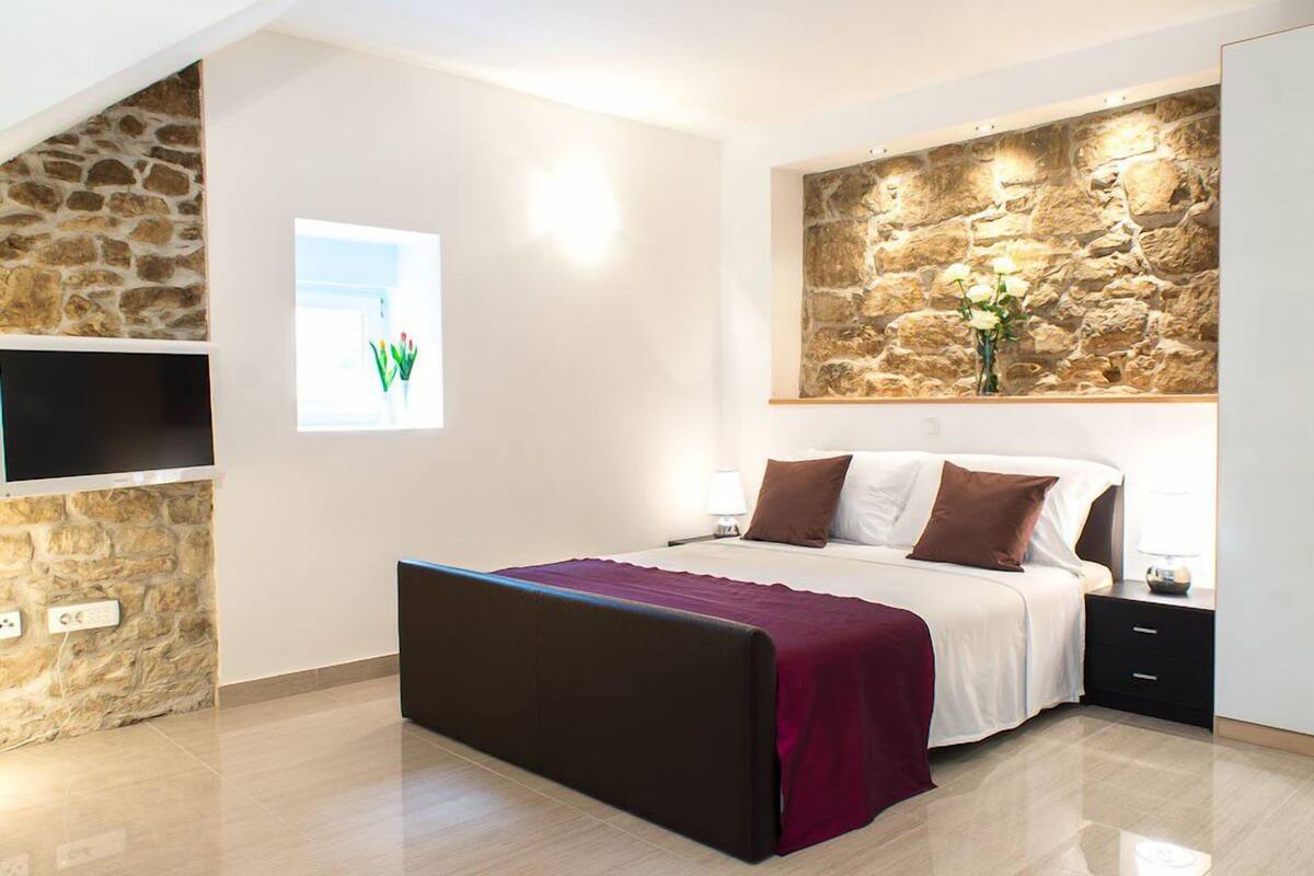 A modern bedroom with a stone accent wall, a large bed with a red throw, a flat-screen TV mounted on a stone wall, and a window with a view of greenery.