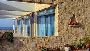 A stone building with a covered patio, large windows with blue curtains, and potted plants. - Aphrodite Beach Hotel (Neo Chorio)