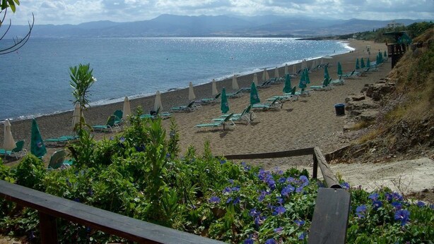 A beach with rows of green lounge chairs and umbrellas, a wooden deck in the foreground, and a view of the sea and distant mountains. - Aphrodite Beach Hotel (Neo Chorio)