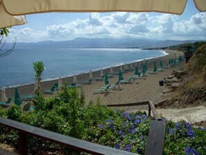 A beach with rows of green lounge chairs and umbrellas, a wooden deck in the foreground, and a view of the sea and distant mountains. - Aphrodite Beach Hotel (Neo Chorio)