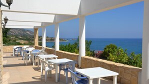 An outdoor patio with white tables and chairs, stone walls, and a view of the sea. - Aphrodite Beach Hotel (Neo Chorio)