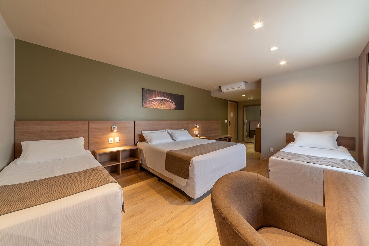 Habitación de hotel con dos camas, un sofá, un cabecero de madera, una obra de arte en la pared y un aire acondicionado instalado en el techo.