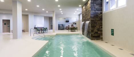 An indoor swimming pool with clear water, surrounded by a tiled floor and walls, with a stone accent wall and a seating area in the background.
