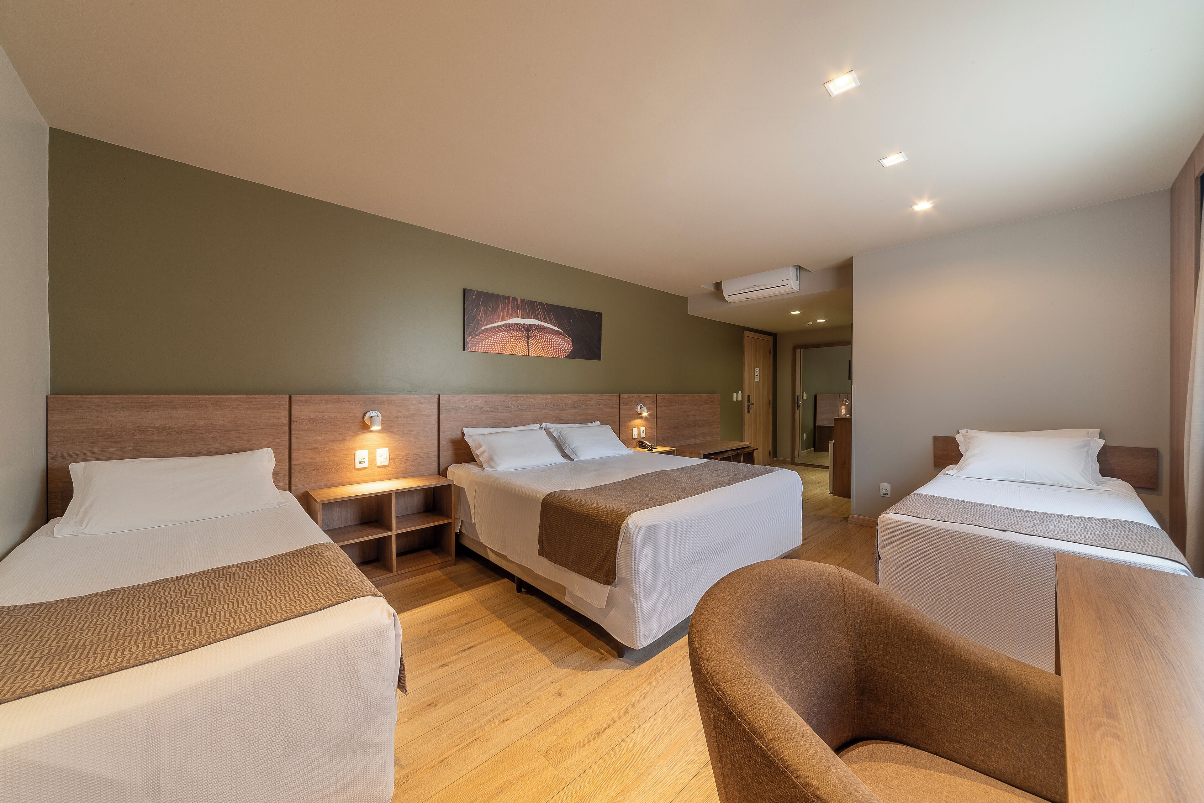 A hotel room with two beds, a wooden headboard, bedside tables with lamps, a wall-mounted air conditioner, and a painting above the beds.