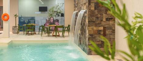 An indoor pool with a waterfall feature, green chairs, and a table with a glass partition.