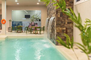 Una piscina interior con una cascada, sillas verdes y una mesa con separador de vidrio.