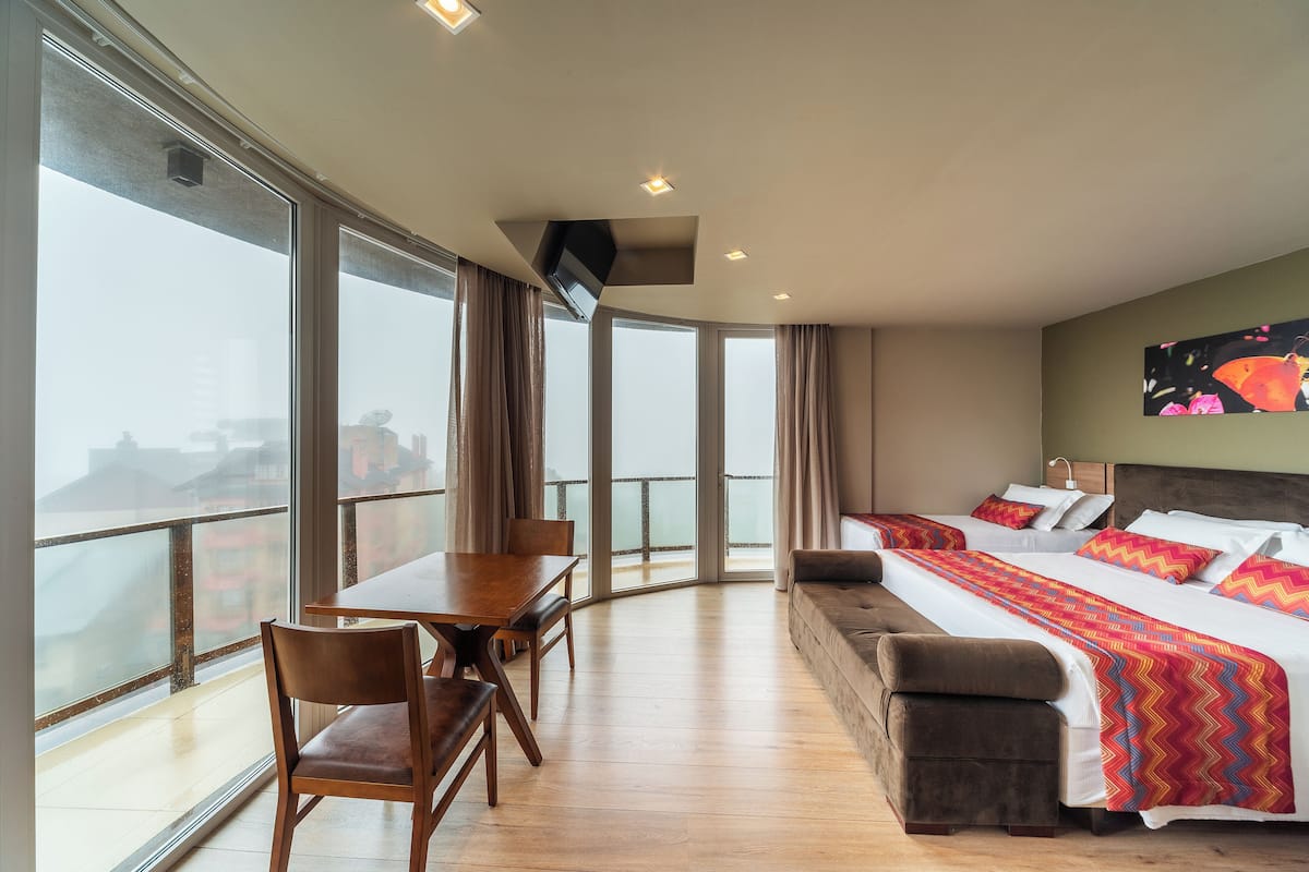 A hotel room with a balcony, a bed with colorful bedding, a sofa, a dining table with chairs, and a TV mounted on the wall.