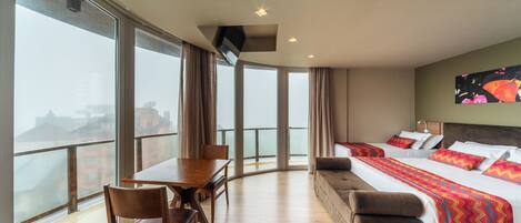A hotel room with a balcony, a bed with colorful bedding, a sofa, a dining table with chairs, and a TV mounted on the wall.