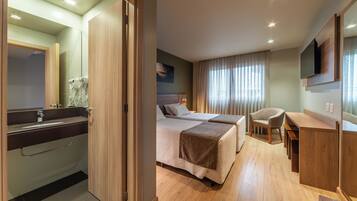 Habitación de hotel con dos camas, un escritorio, una silla, un televisor montado en la pared y un baño con espejo y secador de pelo.