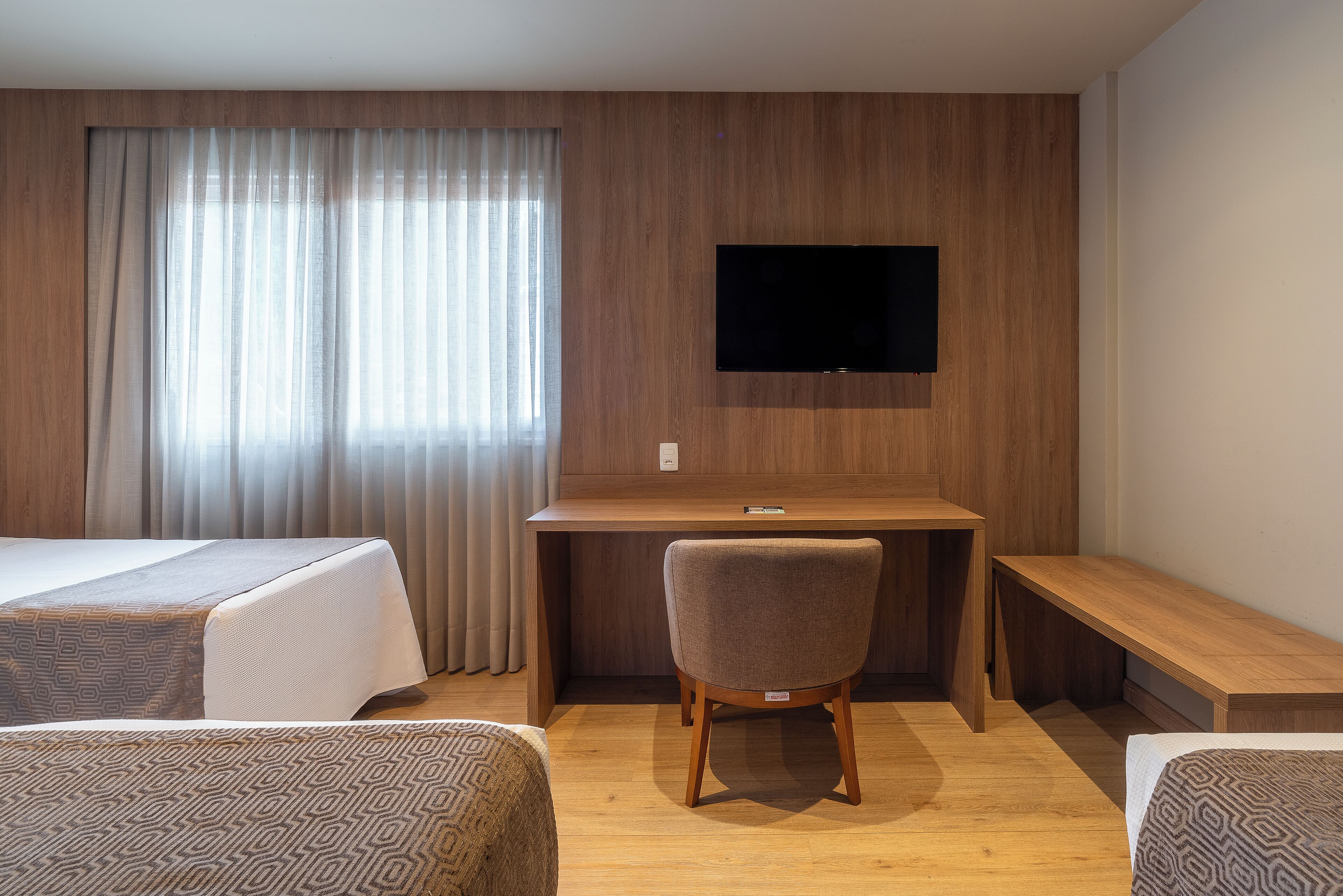 A hotel room with two beds, a desk, a chair, a TV mounted on the wall, and a large window with curtains.