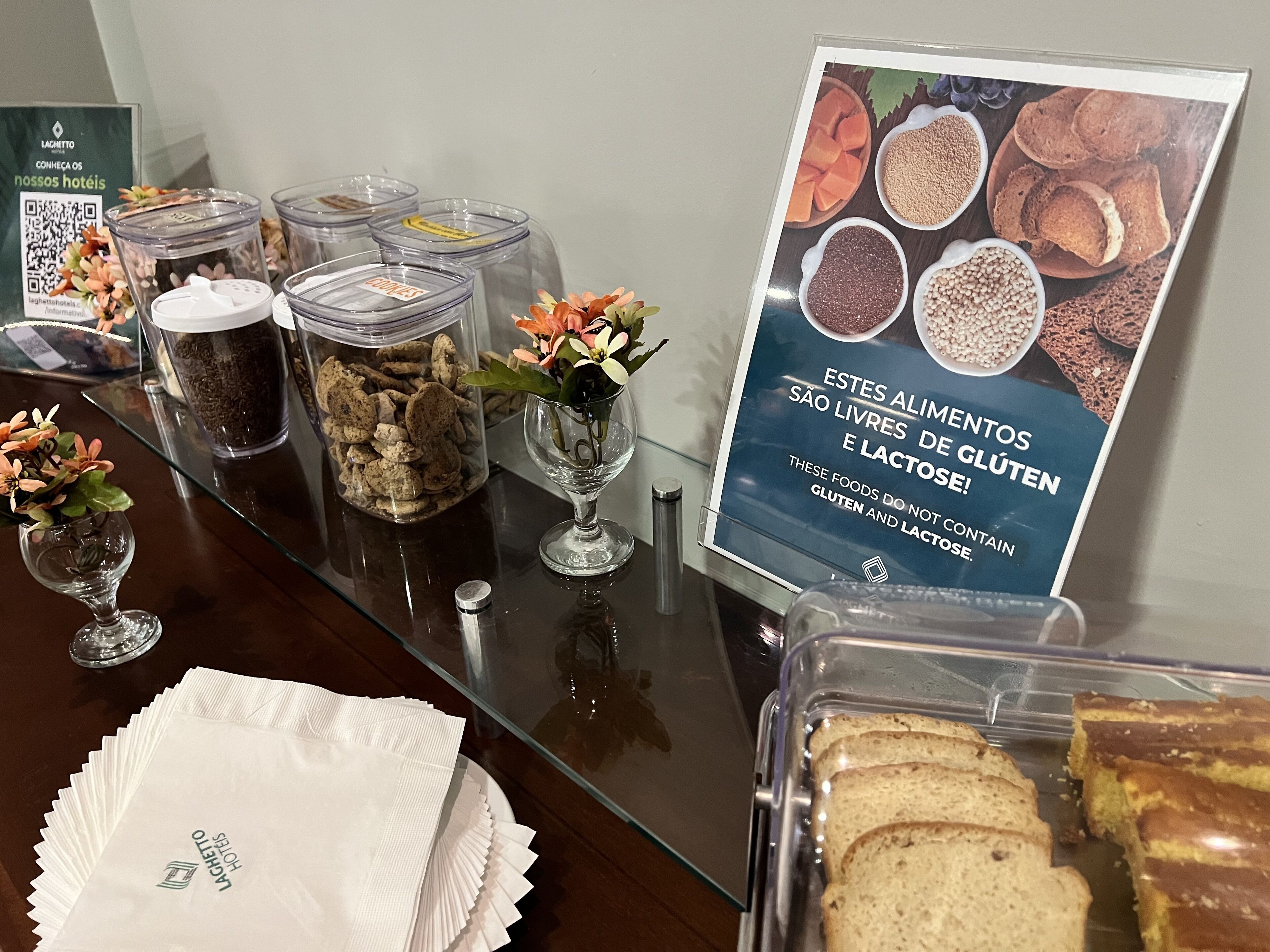 Zona de bufé con diversas opciones de comida, que incluyen cereales y pan, y un cartel que indica opciones sin gluten y sin lactosa.