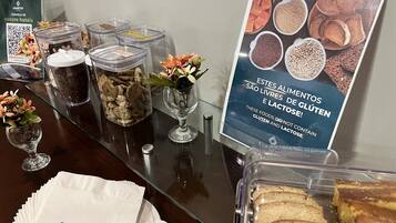 Zona de bufé con diversas opciones de comida, que incluyen cereales y pan, y un cartel que indica opciones sin gluten y sin lactosa.