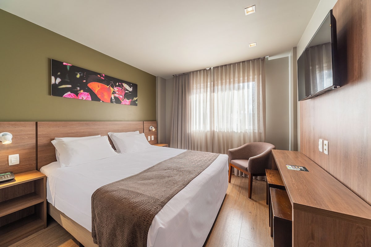 A hotel room with a large bed, a desk, a chair, a TV, and a painting on the wall.