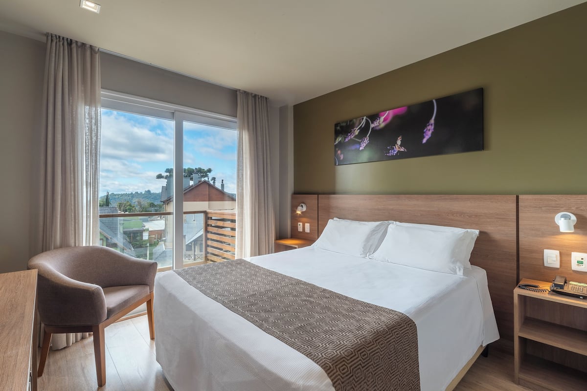 A hotel room with a large bed, a desk, a chair, a window with curtains, a painting on the wall, and a view of buildings outside.