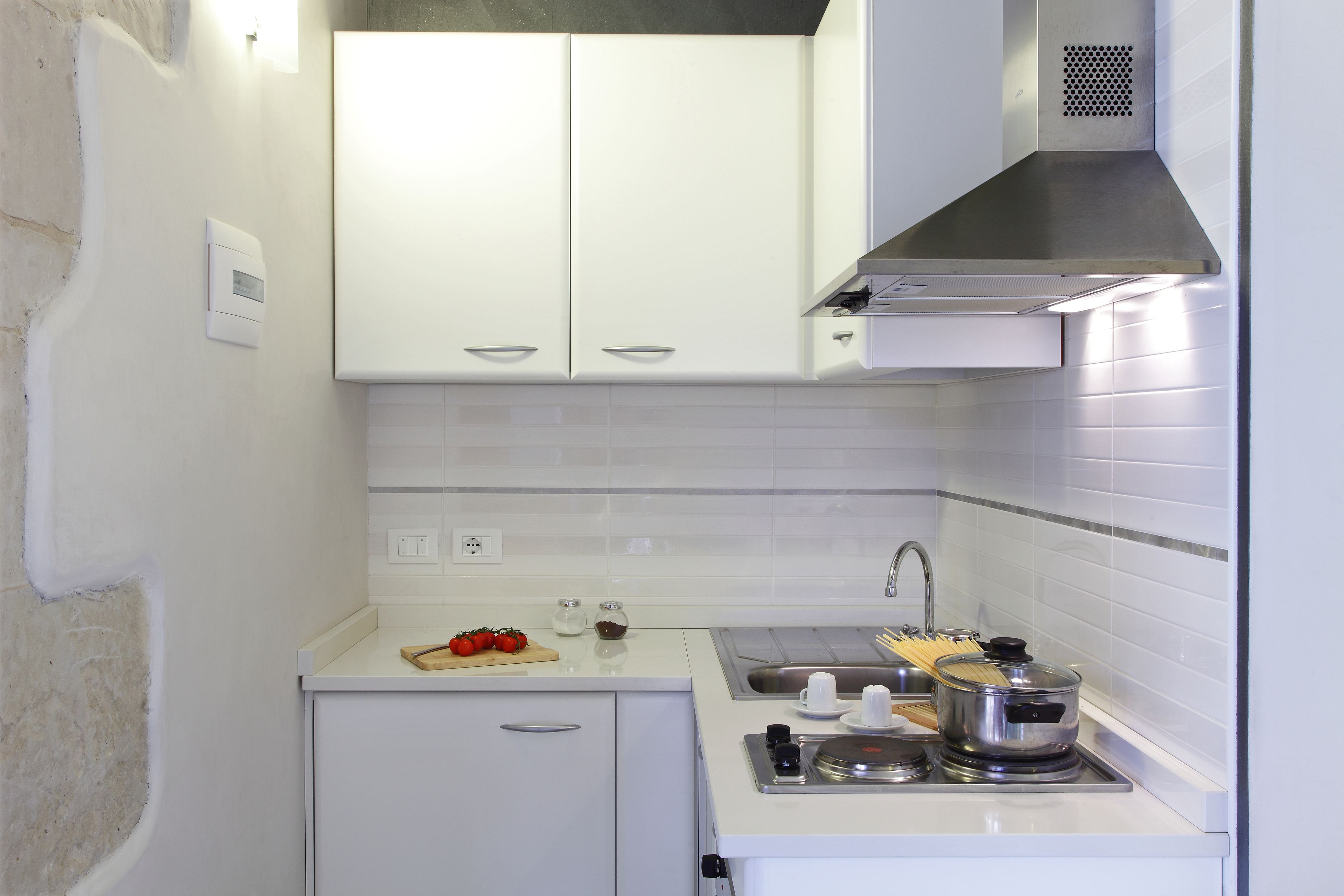 A small kitchen with white cabinets, a stainless steel range hood, a sink, and a countertop with a cutting board, tomatoes, and a pot.