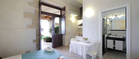 A bedroom with a bed, a table with chairs, a bathroom with a sink and mirror, and a door leading to a balcony.