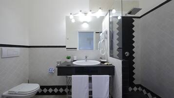 Un baño con inodoro blanco, lavamanos y espejo con iluminación. Hay toallas colgadas en un perchero y una planta pequeña en la encimera.