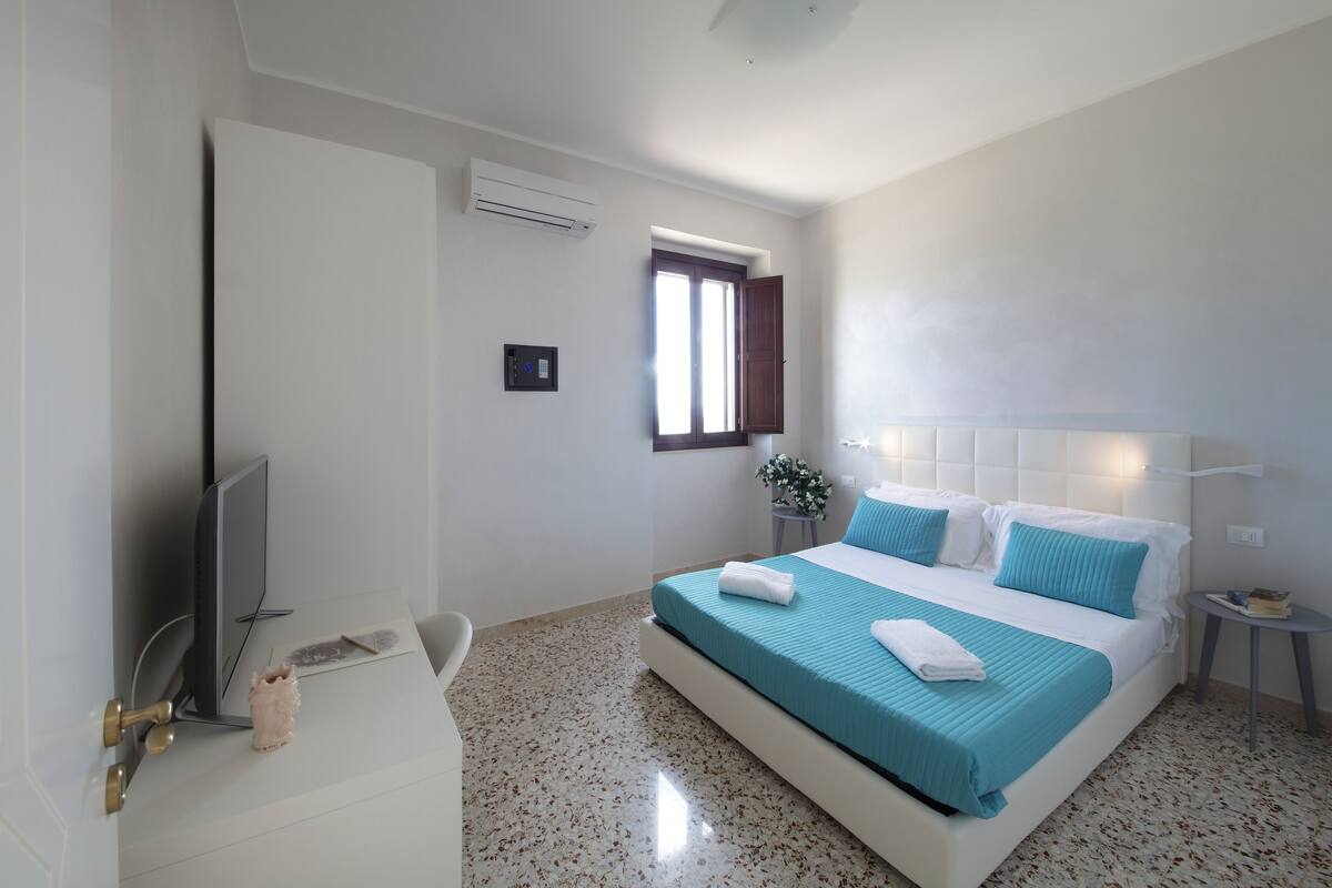 A modern hotel room with a bed, a TV, a desk, and a window.