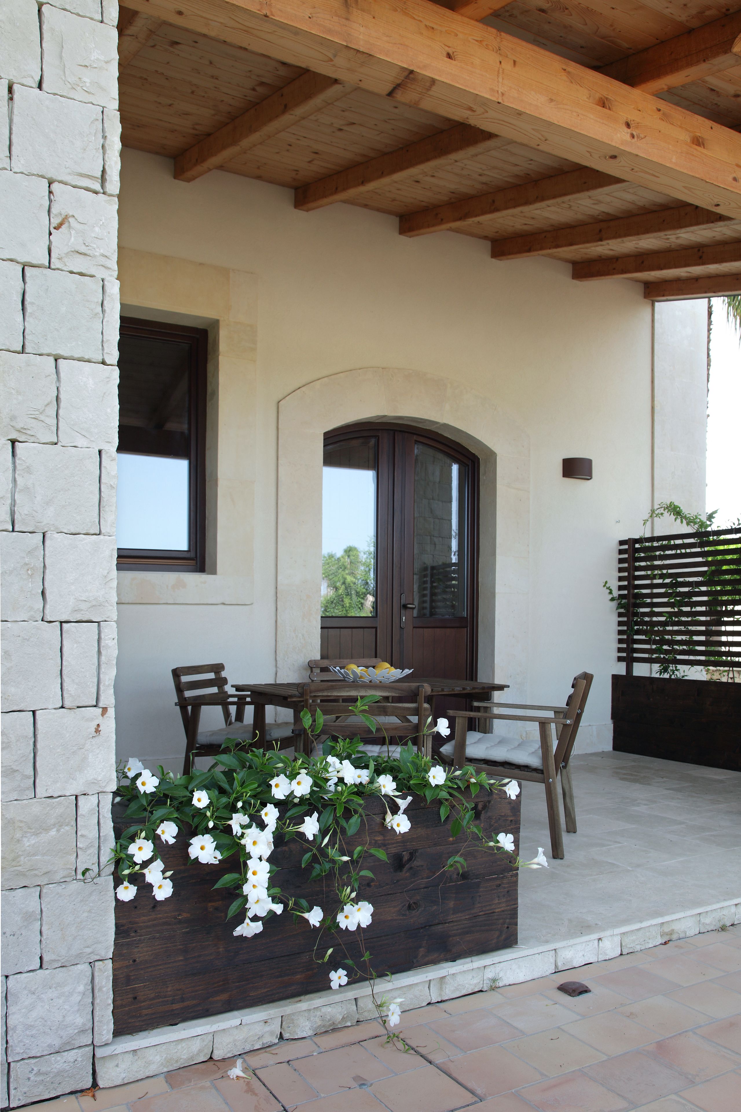 A patio with a wooden table and chairs, a stone wall, and a wooden planter with white flowers.