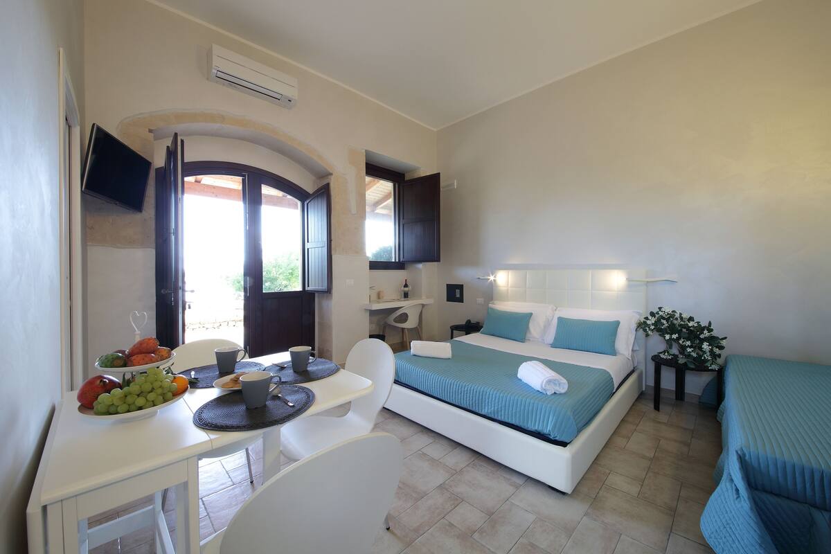 A bedroom with a bed, a desk with a fruit bowl, a chair, and a TV mounted on the wall.