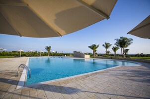 Piscina com borda de pedra, rodeada por guarda-sóis e palmeiras.