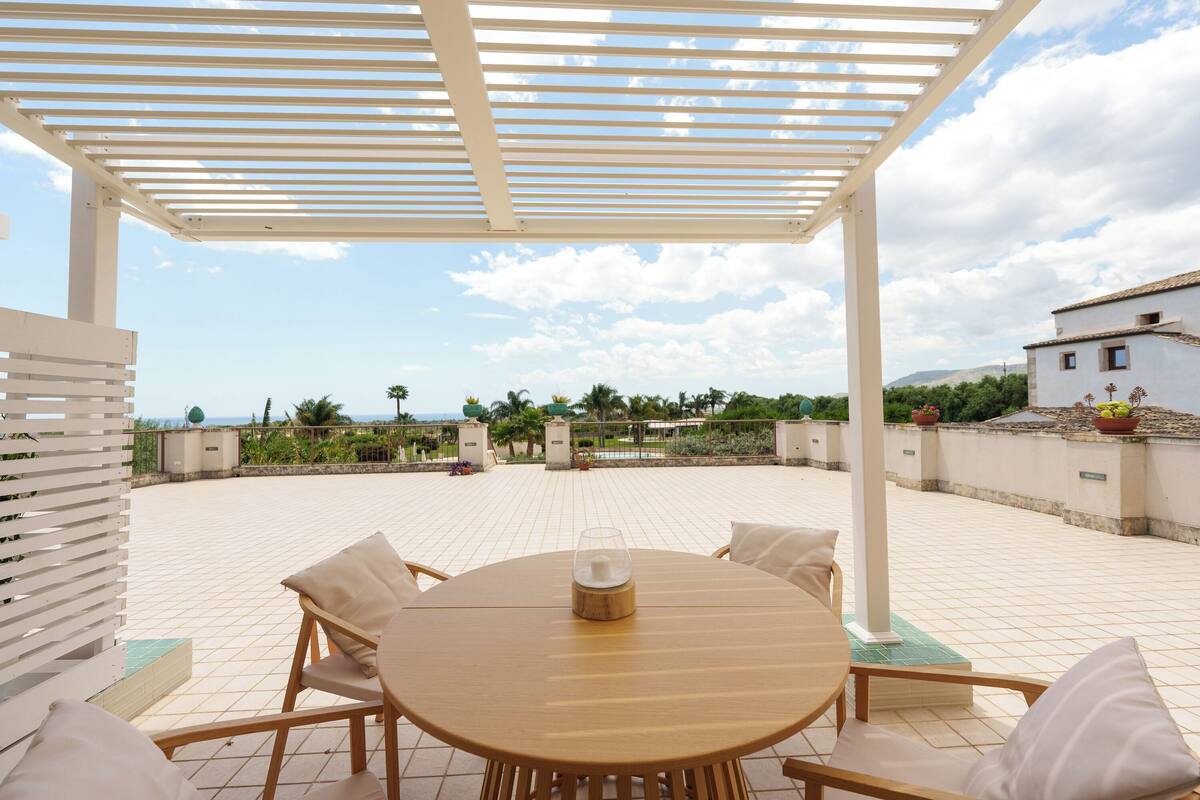 A covered outdoor patio with a round table and chairs, a pergola overhead, and a view of the landscape.
