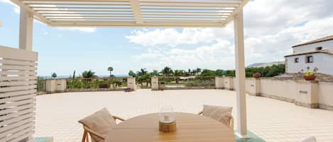 Un patio exterior techado con una mesa redonda y sillas, una pérgola arriba y vistas al paisaje.
