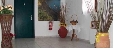 A hallway with a door, a painting of a coastal landscape, a vase with dried reeds, a red vase with flowers, and a small table with a basket.