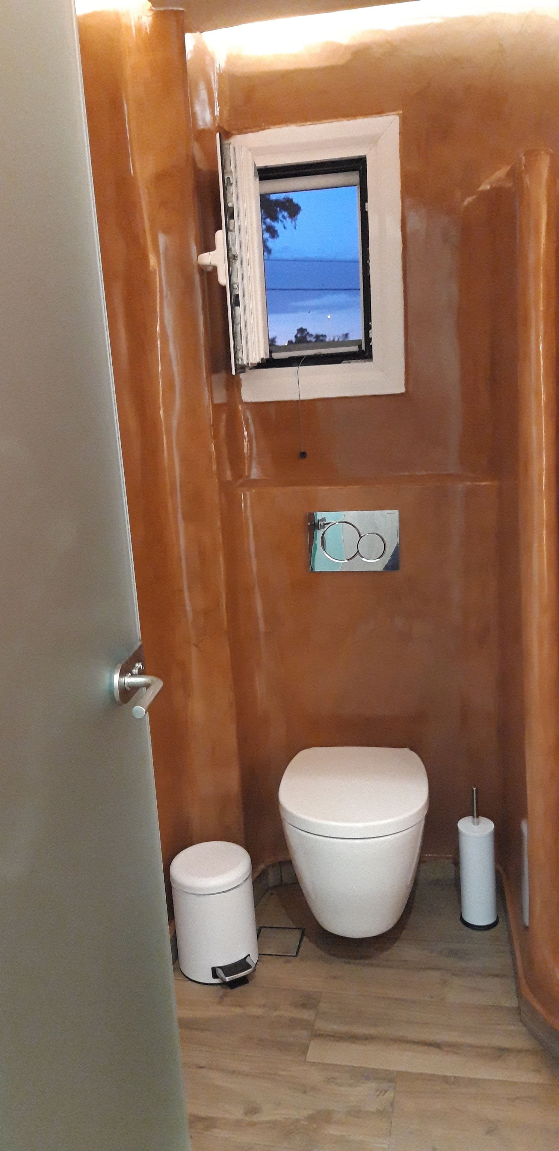 A small bathroom with a white toilet, a small trash bin, and a window with a view of the sea.
