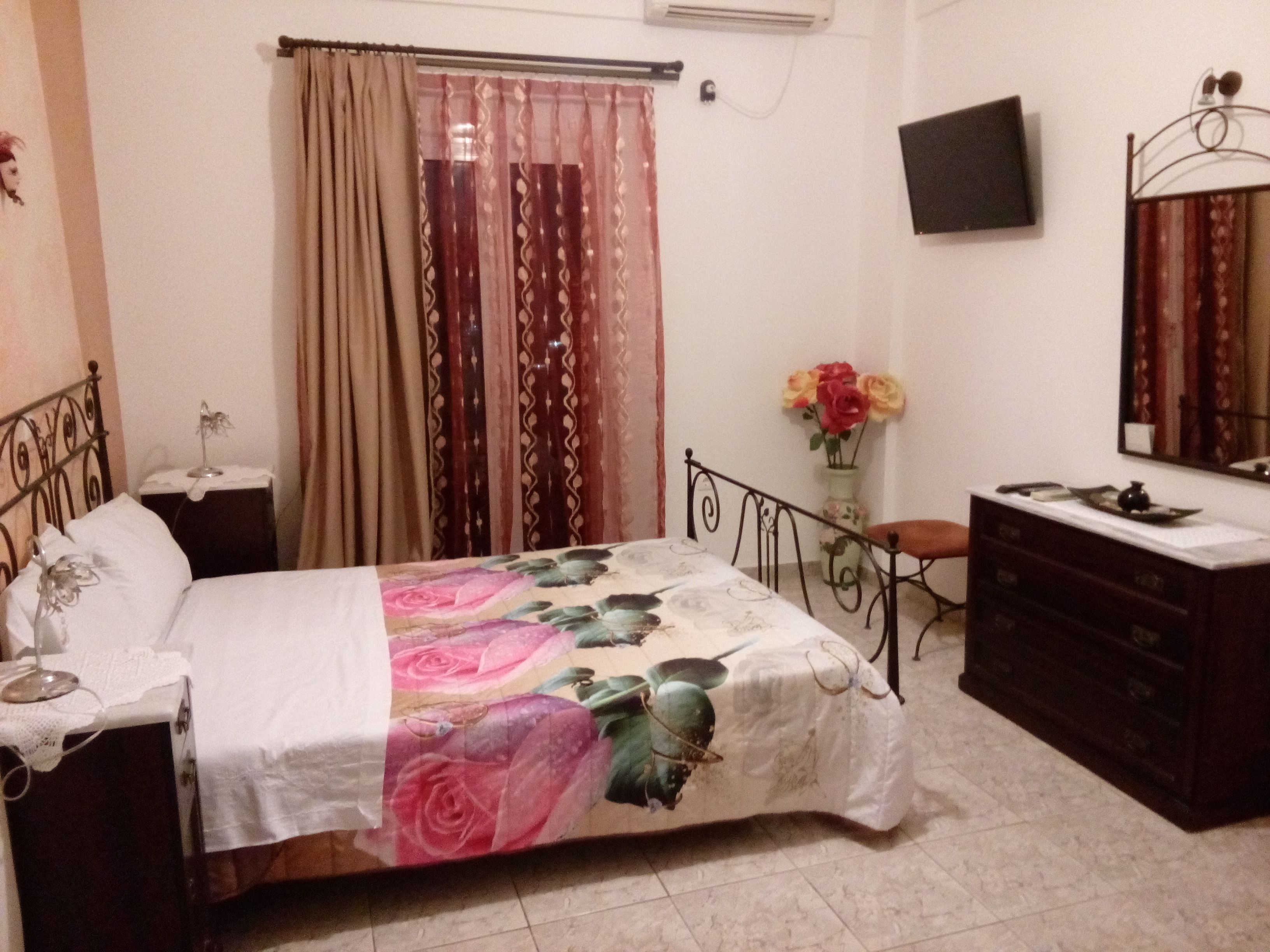 Un dormitorio con cama, cómoda, mesita de noche, espejo, televisor y ventana con cortinas.
