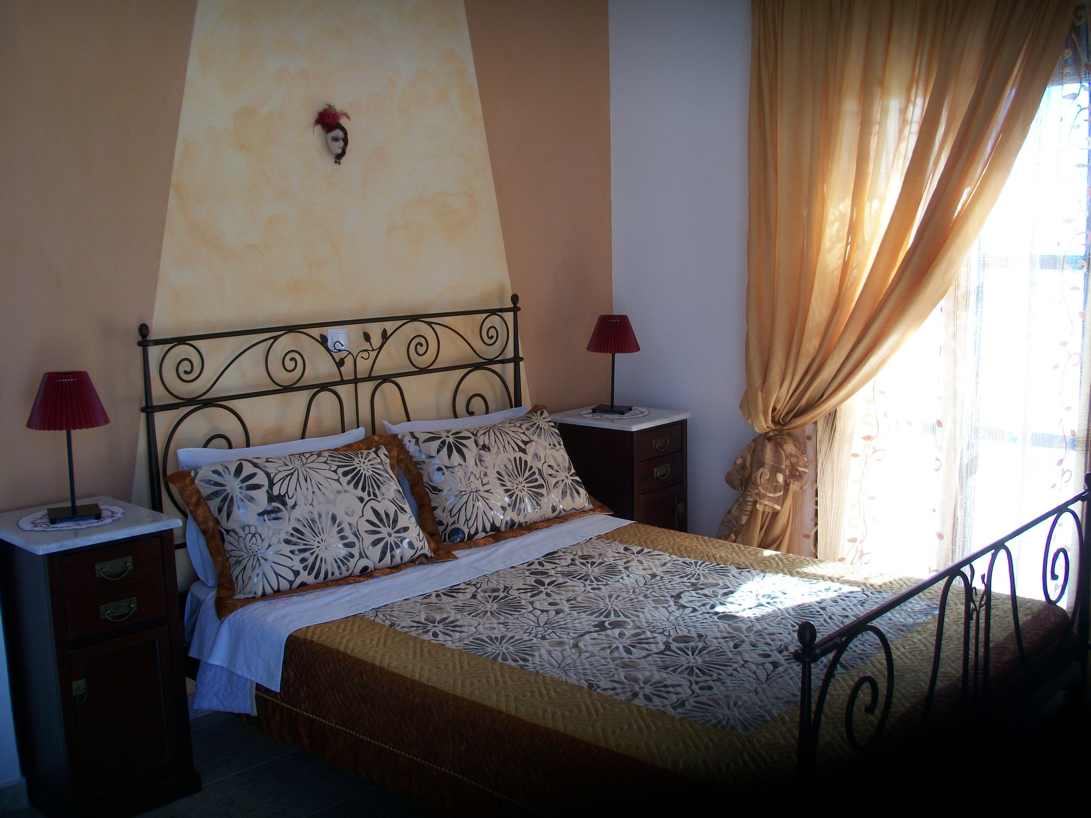 A bed with a patterned bedspread, two bedside tables with lamps, a wall-mounted light fixture, and a window with curtains.