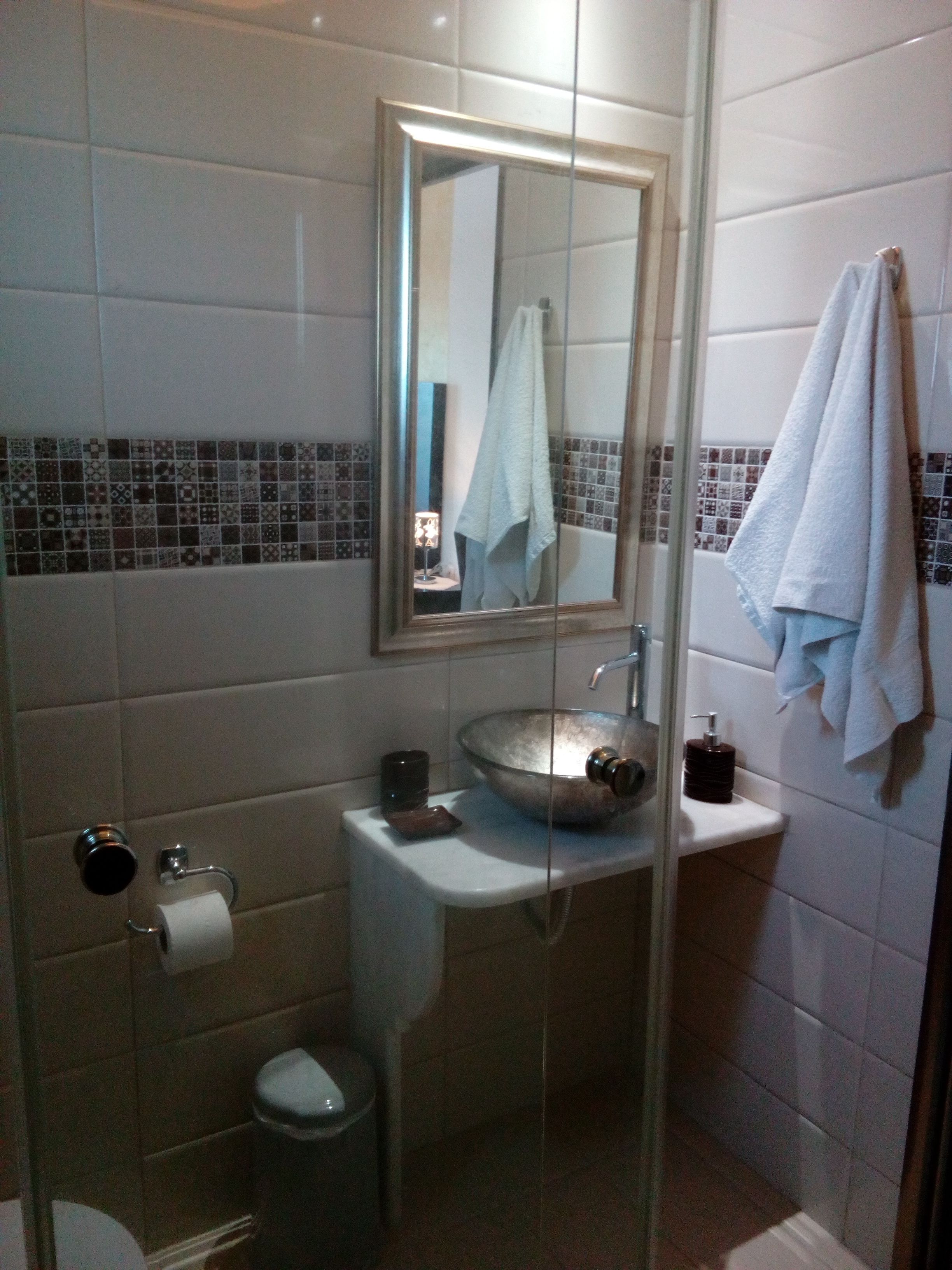 Un baño con ducha envuelta en vidrio, un lavamanos con un recipiente amplio, un inodoro y un espejo sobre el lavamanos.