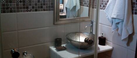 Un baño con ducha envuelta en vidrio, un lavamanos con un recipiente amplio, un inodoro y un espejo sobre el lavamanos.