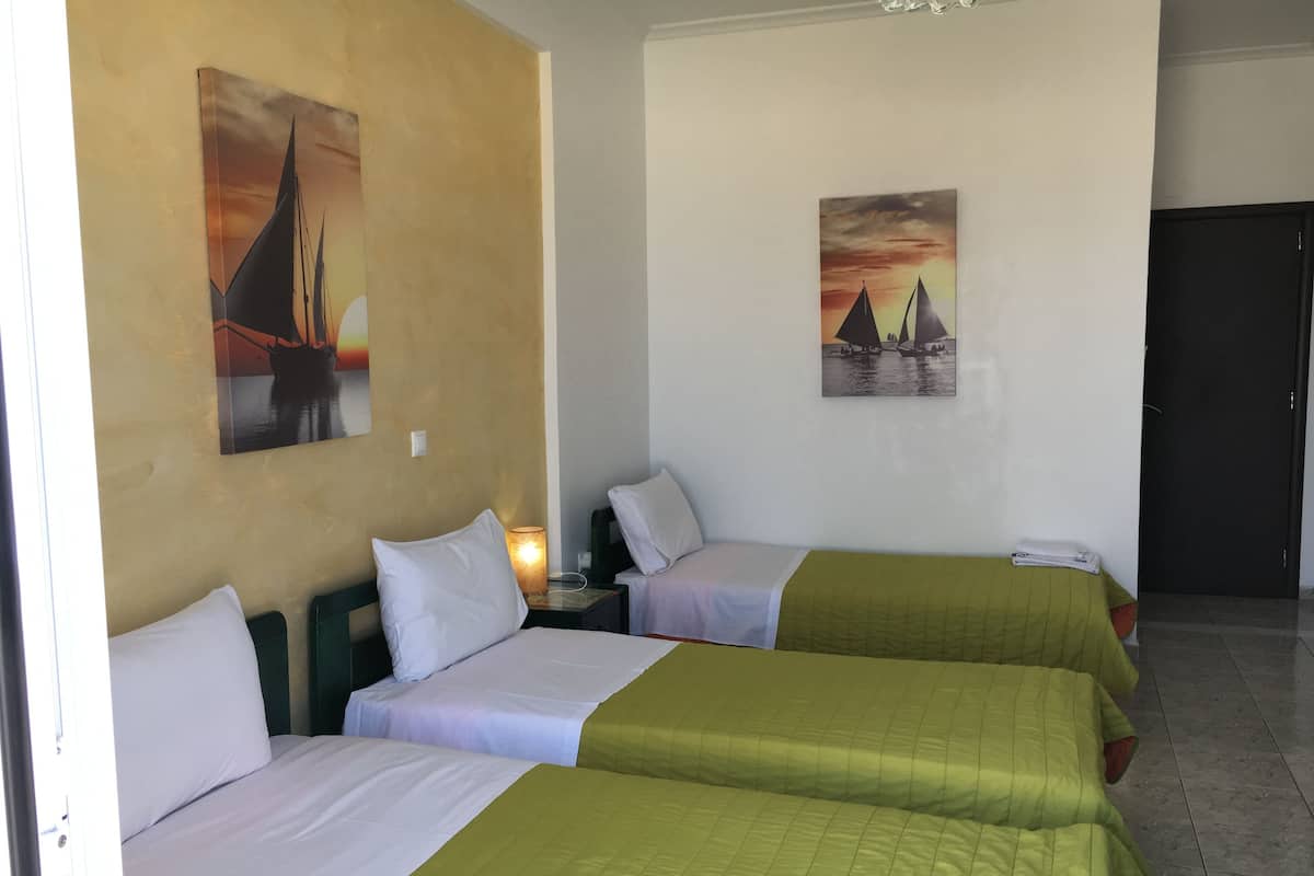 A hotel room with three beds, each with a green bedspread and white linens. There are two paintings on the walls, one depicting a sunset and the other a sailboat.