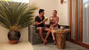 Un hombre y una mujer sentados en un banco, cada uno sosteniendo una bebida de coco. Están a un lado de una planta grande en maceta y una pequeña mesa de madera.