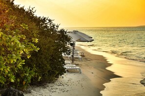 Ein Strand mit weißen Liegestühlen und Sonnenschirmen, einem Sandweg und dichter Vegetation.