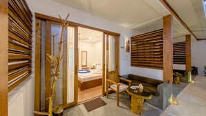 Una habitación con una cama, persianas de madera, una silla, una mesita y una planta decorativa.