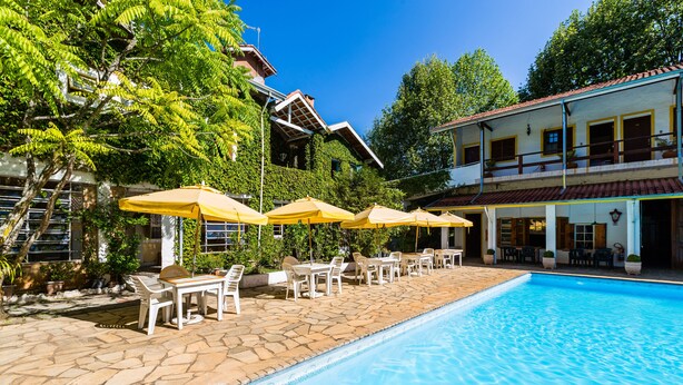 A swimming pool with stone tiles, surrounded by white lounge chairs and yellow umbrellas, and a building with a red roof and balconies. - Pousada Cesar (São Bento do Sapucaí)