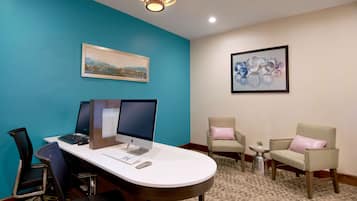 A conference room with a desk, two monitors, a keyboard, and a mouse. There are two chairs with cushions, a small table with a plant, and a framed picture on the wall.