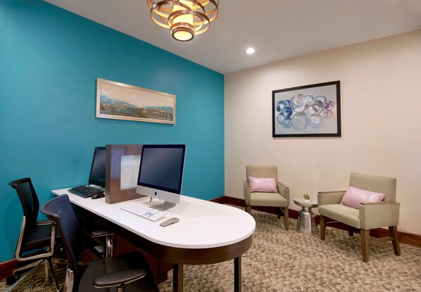 A conference room with a desk, two monitors, a keyboard, and a mouse. There are two chairs with cushions, a small table with a plant, and a framed picture on the wall.