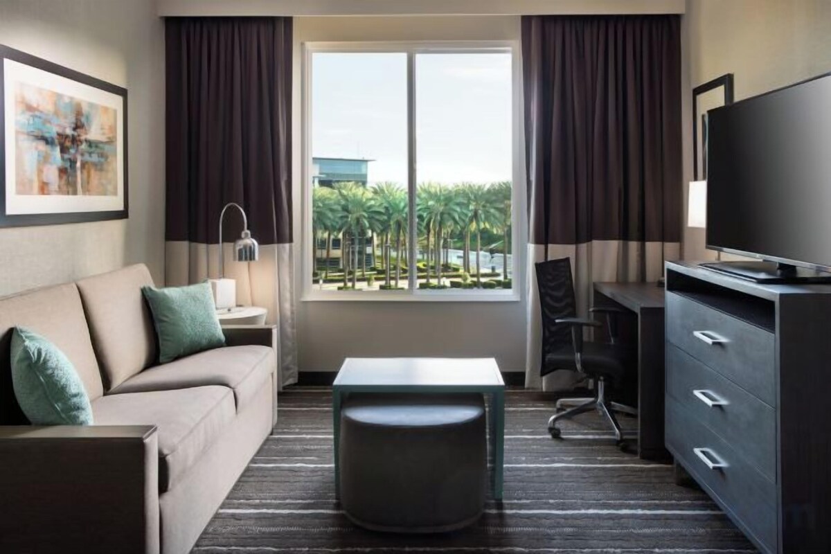 A hotel room with a sofa, a small table, a TV, and a view of palm trees.