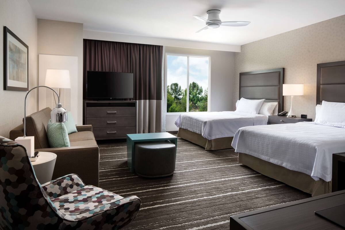 A hotel room with two beds, a TV, a desk, and a view of the outdoors.