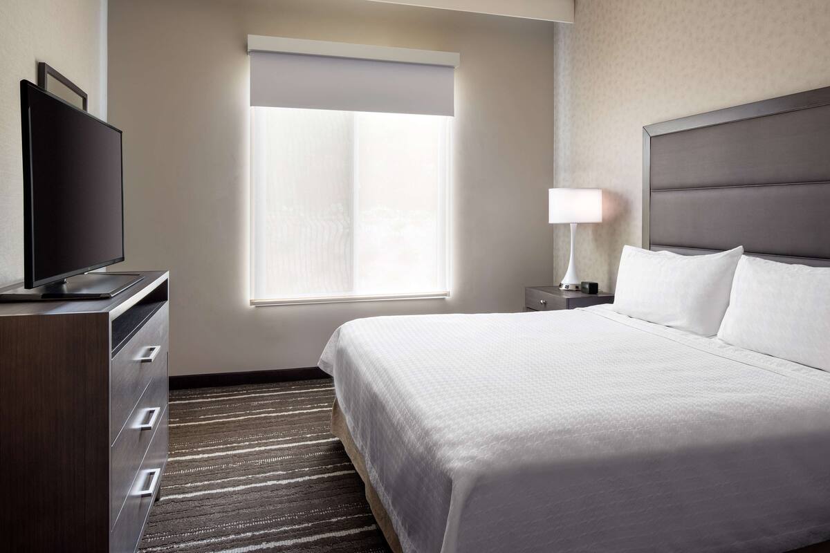 A hotel room with a bed, a television, a nightstand, a lamp, and a window with blinds.