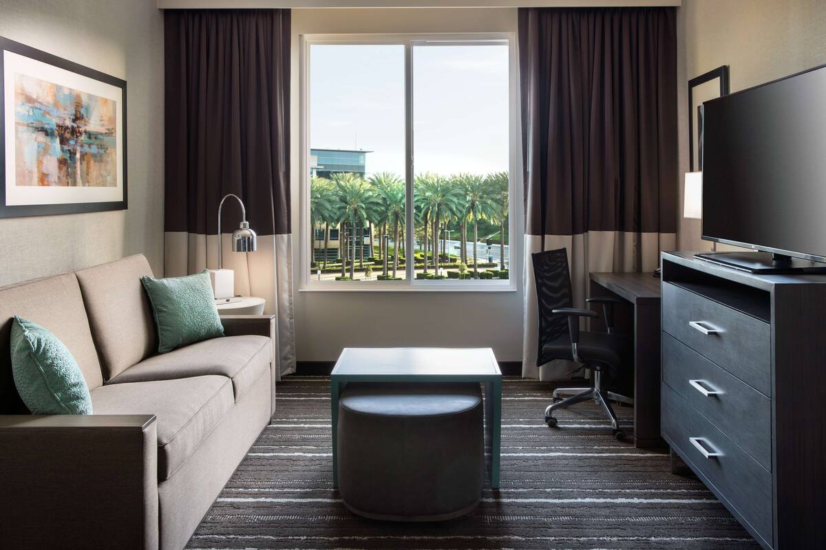 A hotel room with a sofa, a small table, a TV, a desk, and a window with a view of palm trees and buildings.