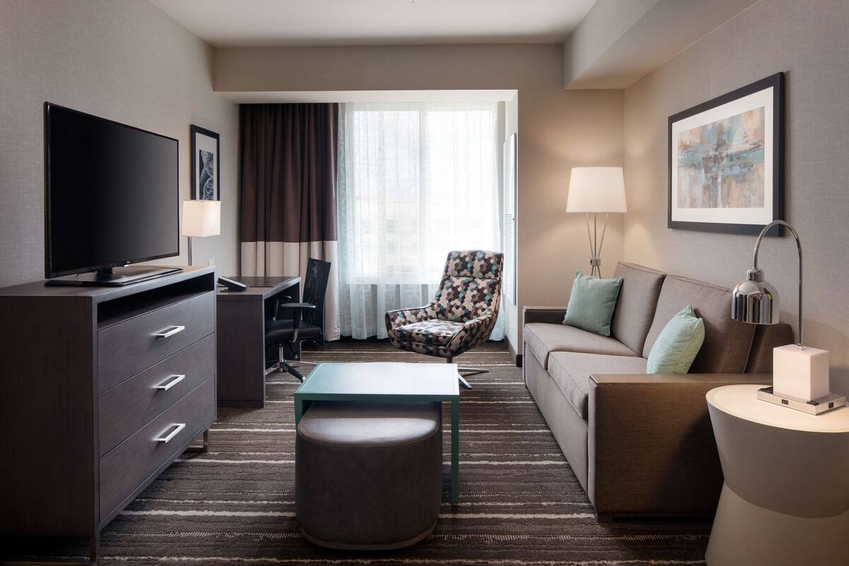 A hotel room with a flat-screen TV, a desk, a patterned armchair, a sofa, a small ottoman, and a side table with a lamp.