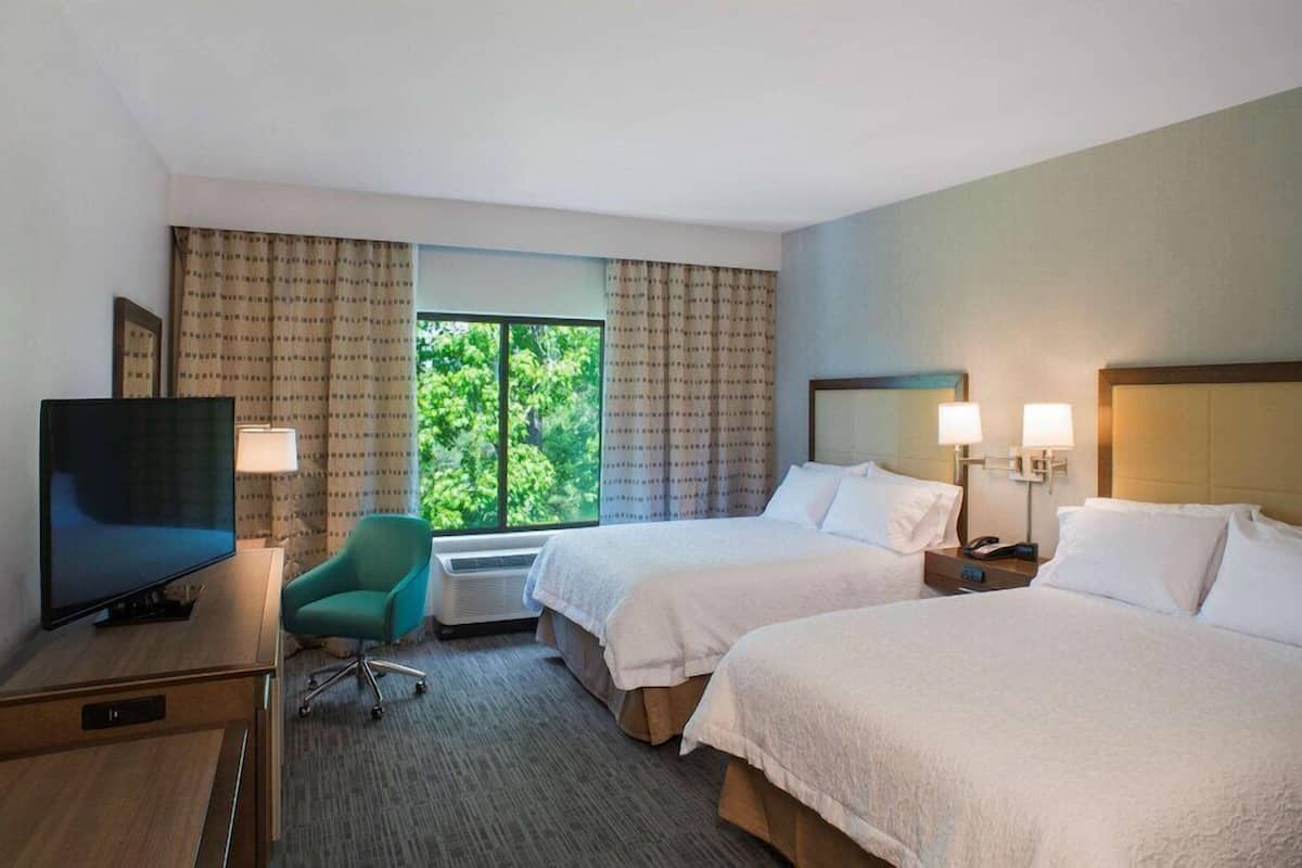 A hotel room with two beds, a desk with a TV, a chair, and a window with curtains.